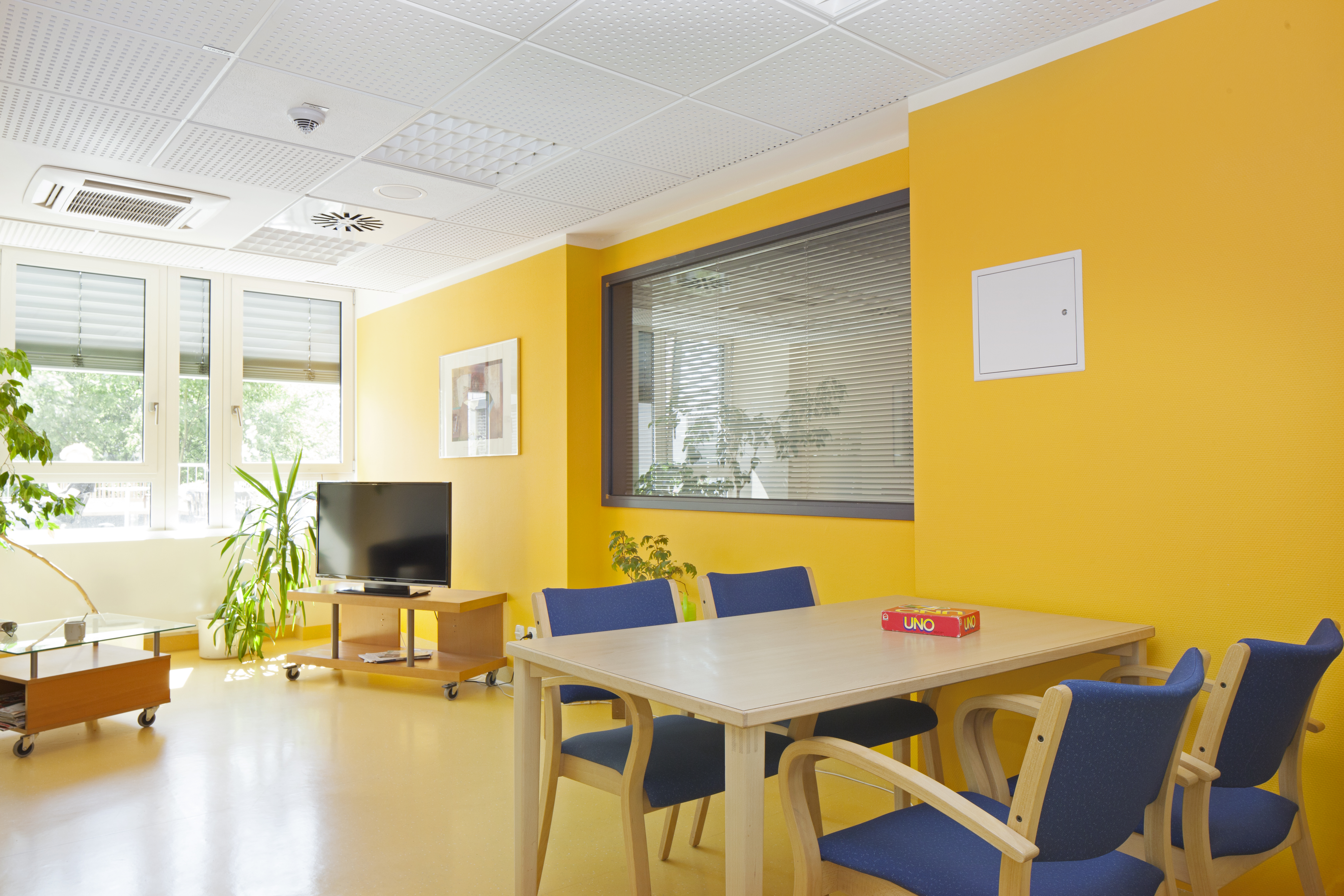 Aufenthaltsraum für Patientinnen und Patienten der Station P1 der LVR-Klinik für Psychiatrie und Psychotherapie Essen mit gelben Wänden, einem Tisch mit vier Stühlen im Vordergrund. Im Hintergrund sind vor den Fenstern ein Fernsehgerät und Pflanzen zu sehen.