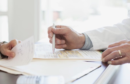 In diesem Bild sieht man eine Hand eines Arztes, die einen Stift hält, auf dem Tisch liegen Unterlagen. Links sitzt eine Person. Es soll ein Arzt-Patienten-Gespräch symbolisieren.